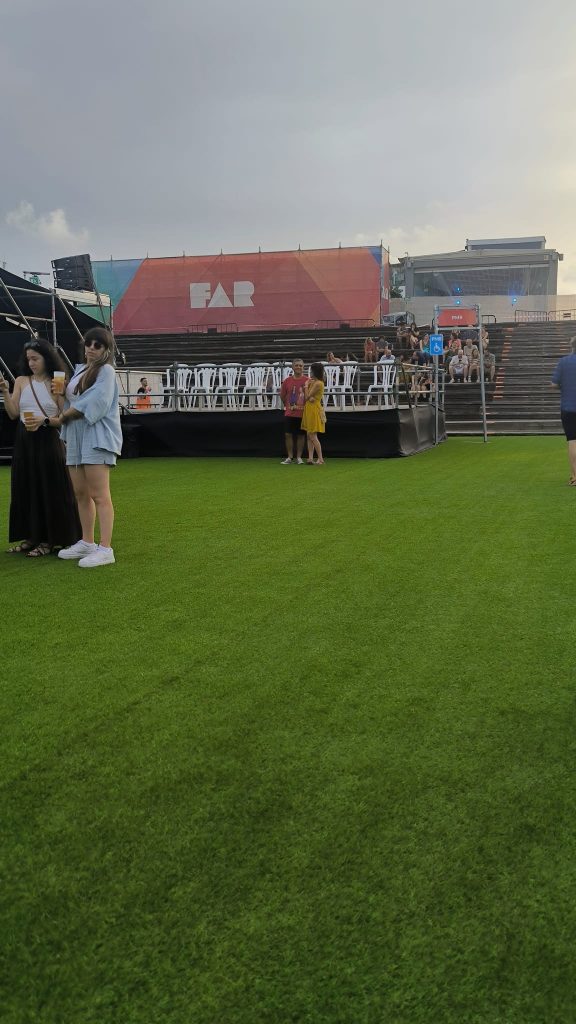 Vista general de la zona de conciertos del Festival FAR. En primer plano se observa la plataforma reservada para personas con movilidad reducida (PMR), elevada sobre el césped artificial y equipada con sillas blancas. Al fondo aparece el escenario principal con la imagen del festival y, junto a la plataforma, la señalización de acceso PMR. La fotografía muestra la ubicación de este espacio, situado junto a la torre de control de sonido, con buena visibilidad del escenario y un acceso específico para las personas usuarias.