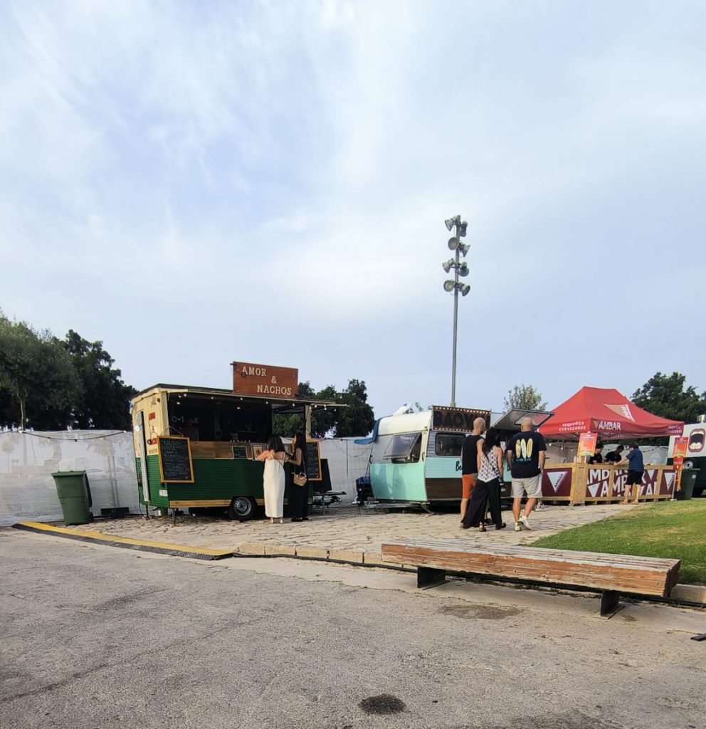 Vista de la zona de restauración del Festival FAR. En primer plano se observa un amplio espacio asfaltado que da acceso a varios food trucks y barras de bebidas situados sobre una plataforma pavimentada con un pequeño desnivel respecto al vial. A la derecha hay una zona de césped y varias personas haciendo cola o caminando entre los puestos. La imagen ilustra el entorno donde se encontraban los espacios de restauración,