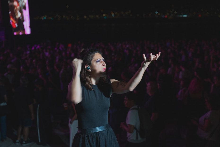 Fotografía nocturna de una intérprete de lengua de signos durante un concierto del Festival Noroeste, con el público al fondo. Alba, vestida de negro, aparece en plena actuación, con ambos brazos elevados mientras signa.